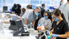 Dental students wearing masks practice procedures on mannequins in a clinical lab with instructors supervising.