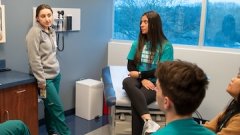 Students in scrubs gathered in a medical exam room, listening to a peer speak during a simulation or training session.