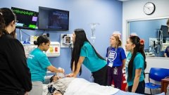 Students in teal shirts observe a medical simulation on a patient mannequin in a hospital training room.
