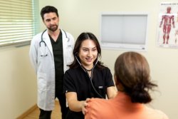 A student uses a stethoscope to examine a patient while a healthcare instructor looks on.