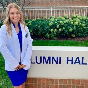 Anja Goetten posing in white coat outside academic building sign.