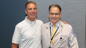 Mr. Giglio and Dr. Borelli in a white coat and stethoscope take a photo together at the patient panel.  