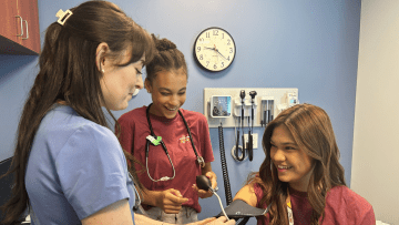 Two high school students learn how to take vital signs from a Midwestern student in the simulation exam room 