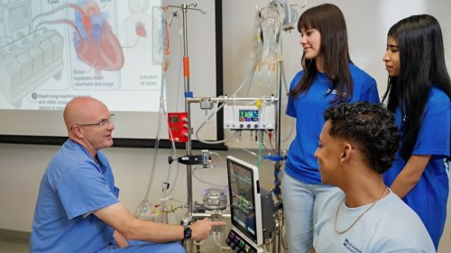 Instructor demonstrates a medical heart-lung machine to a small group of students in a clinical training lab with monitors and tubing visible.