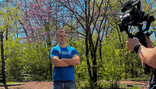 Daniel Misioura stands among trees with a cameraman at Midwestern University.