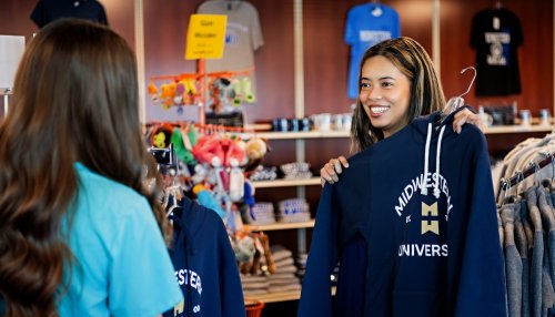 A shopper smiles while holding up a Midwestern University hoodie to show a friend inside a colorful campus bookstore.