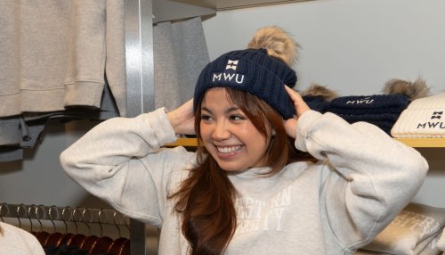 A smiling student tries on a navy knit beanie with an MWU logo inside a campus store, surrounded by folded apparel.