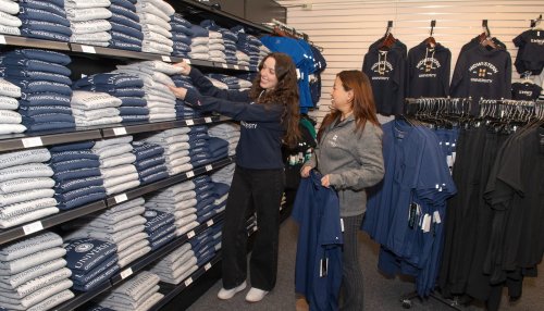 Two shoppers browse stacks of folded Midwestern University branded sweatshirts and hanging apparel inside the campus store, smiling as they select clothing.