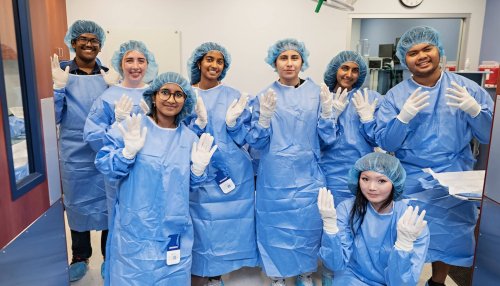 Eight Health Careers Institute high school students in blue surgical gowns, gloves, and hair caps pose together in a clinical training room at MWU's Glendale campus.