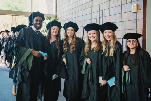 Six graduates take a group photo in a line with black caps and gowns and carrying their name cards.