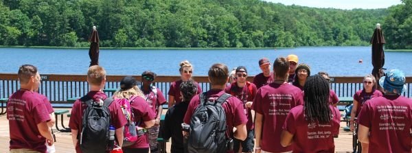 Attendees gather in a group by the lake at the EDI camp wearing burgundy camp shirts.