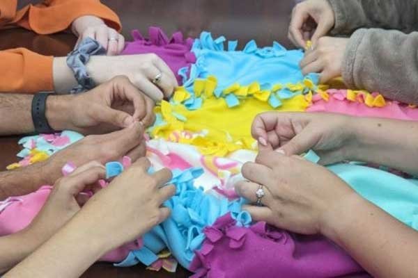 Students work together to make no-sew blankets. Five pairs of hands make the no-sew blankets in pink, yellow, turquoise, and purple material.