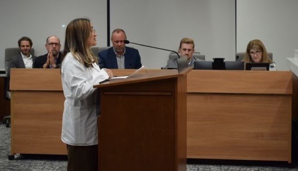 Bethany Jimenez delivers a speech at a podium during the Downers Grove Village Council Meeting in a white coat.