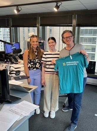 Students Tiffany Thompson and Alexis Flamburis with WGN host John Williams in the WGN Studio. John WIlliams holds a teal Midwestern University T-shirt.