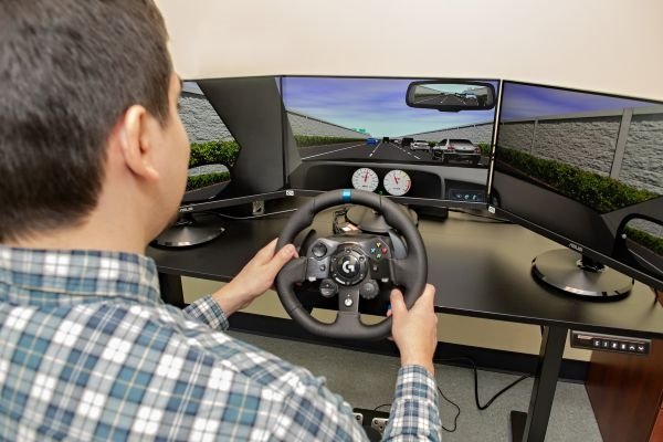 A participant demonstrates the driving simulator holding the wheel, navigating traffic, with a realistic driving scenario on the screens.